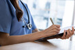 © 220 Selfmade studio - Focused female medical professional wearing blue scrub and stethoscope writing patiently on patient clipboard chart working diligently in bright hospital room setting