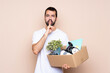 © luismolinero - Man holding a box and moving in new home over isolated background showing a sign of silence gesture putting finger in mouth