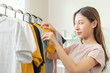 © KMPZZZ - Choice of clothes,Nothing to wear. Attractive asian young woman, girl try on appare, happy choosing dress, outfit on hanger in wardrobe in room closet at home. Deciding blouse what to put on which one