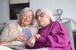 © luciano - Smiling couple of senior women sitting together on couch sharing technology and social media by smartphone while having pleasant conversation. Friendship and companion in elderly retirement life