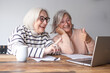© luciano - Two smiling senior women with white hair and glasses sitting at a wooden table at home, looking at a laptop together. They appear relaxed and happy while browsing online, with papers and a coffee mug