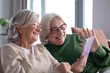 © luciano - Cheerful couple of senior women sitting together on couch sharing technology and social media by smartphone while having pleasant conversation. Friendship and companion in elderly retirement life
