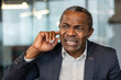 © Liubomir - Mature man grimacing, inserting finger into his ear, indicating an irritating noise, a sudden loud sound, or experiencing an earache, tinnitus, or hearing issues at work