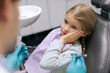 © Home-stock - Child girl sitting in dental chair having pain in tooth, touching cheek and looking at male dentist