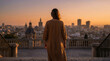 © Video80X - A woman in a brown dress stands with her back to the camera, gazing at a city skyline during sunset from a stone terrace with railings.