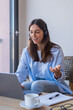 © Daniel - Friendly young woman wearing a headset engages in a remote video meeting from a bright home workspace, using a laptop with notes and coffee nearby, conveying communication, professionalism and ease.