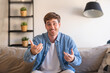 © Daniel - Smiling young man sits on a couch in a denim shirt, gesturing warmly during a friendly video call or conversation at home, conveying approachability, communication and a relaxed modern lifestyle.