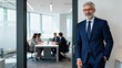 © ibhonk - Mature businessman with gray hair and glasses stands confidently in a modern office doorway while a team works in the background.