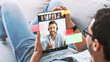 © Prostock-studio - A young man with glasses lies on a sofa looking at a tablet. He is participating in a video call while searching for job opportunities. The setting is casual and relaxed.