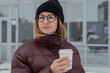 © Alexander - Woman sipping coffee outside storefront, casual streetstyle portrait with confident expression, maroon puffer coat and black beanie, round glasses catching soft light, neutral winter tones, subtle