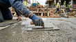 © Ahsan - A construction worker kneels, using a trowel with a wooden handle to spread and smooth fresh concrete on a building foundation.