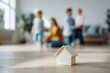 © Andrii Zastrozhnov - Small wooden house toy in foreground with blurred caucasian family male female adults and young children in cozy living room