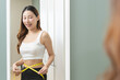 © KMPZZZ - Weight lose, loss concept. Slim body asian young woman hand use tape measuring around waistline in fit sports. Girl looking reflect in mirror at home. Healthy nutrition, fitness for wellbeing beauty.