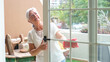 © luciano - Elderly woman with glasses cleaning a glass door with a pink cloth in a bright modern home interior, representing household chores and everyday domestic life