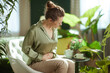 © Alliance - Surrounded by houseplants, a woman in a green silk blouse holds her abdomen, focusing on nervous system regulation and mindful rest during a moment of physical pain.