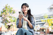 © bongkarn - Pretty asian woman looking at phone as listening to music while sitting on bench under warm sunlight
