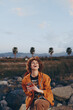 © SHOTPRIME STUDIO - Happy woman with curly hair wearing orange jacket laughs holding smartphone outdoors near rocks with palm trees and mountains in background during casual lifestyle moment.