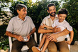 © kleberpicui - Grandfather, adult son and young grandson sitting outdoors and interacting with affection, representing family bonding and multigenerational connection.