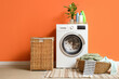 © Pixel-Shot - Modern washing machine with bottles of detergent and laundry baskets near orange wall