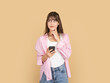 © Designerant - Thoughtful young woman holding smartphone and touching chin, wearing pink shirt and denim, standing against beige studio background with copy space. Thinking and decision concept.