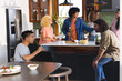 © wavebreak3 - Diverse friends chatting and holding drinks in modern home kitchen around white island with chips