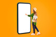 © Prostock-studio - A person stands next to a tall smartphone on an orange background. She touches the screen with one hand and wears a hat and backpack. Sunlight brightens the scene.