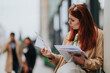© qunica.com - Red-haired business worker reads papers and holds a folder while standing outdoors. A professional woman reviews documents and looks focused on the page.