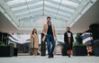 © qunica.com - Three people walk confidently through a modern atrium carrying bags after a business meeting. Colleagues stride together in an urban office and shopping complex, projecting teamwork and confidence.