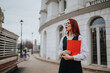 © qunica.com - A professional woman with red hair and glasses holds a red clipboard outdoors. She stands beside a white modern building on a city street, projecting a confident, businesslike mood.