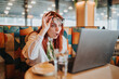 © qunica.com - A surprised businesswoman lifts her glasses and looks at her laptop screen in a cafe. She reacts with shock while working remotely at a table in a colorful booth.