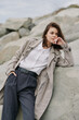 © SHOTPRIME STUDIO - Young woman in textured coat and white shirt leans on rock outdoors, looking sideways with thoughtful expression. Casual fashion, nature, and relaxed mood in natural light.