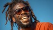 © Jojo* - Close-up portrait of a joyful Black man with dreadlocks and sunglasses laughing outdoors against a clear blue sky.