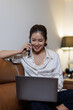 © David - In this image, a smiling woman is engaged in a phone conversation while comfortably using her laptop, showcasing the joy of multitasking in a stylish home environment.