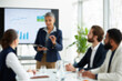 © REAL  - soft focus blurred photo of Professional business presentation in modern office, mature female leader speaking to diverse team, corporate meeting with data charts on screen