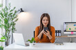 © Tj - Asian businesswoman in a modern office, smiling and focused while checking her smartphone and managing social media, working remotely on a laptop at a clean desk