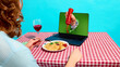 © master1305 - Hand squeezing ketchup from laptop screen onto pasta plate on checkered table with wine glass. Concept of online food delivery advertising, digital dining experience, virtual service promotion.