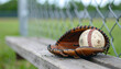 © hardvicore - Baseball leather glove with ball on wooden bench, blurred chain link fence and green field in background, sport and game preparation