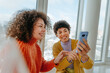 © Westend61 - Smiling friends with curly hair talking and using phone by window