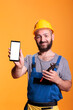 © DC Studio - Construction worker holding cell phone with empty screen for advertising in front of camera. Professional builder with hard hat against yellow background in studio shot.