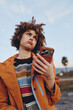 © SHOTPRIME STUDIO - A young woman with curly hair wearing a striped rainbow sweater and orange jacket holds a smartphone outdoors. The woman looks confused or frustrated under a clear sky.