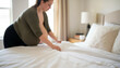 © Aline Shawn - A woman is making a bed, smoothing out the white sheets in a softly lit bedroom.