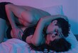 © los3amigos - Una pareja se besa apasionadamente sobre una cama blanca iluminada con luces de neón.