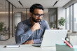 © insta_photos - Young serious indian professional business man, focused ethnic male student wearing glasses working on laptop, remote studying using computer looking at screen watching seminar webinar at home office.
