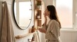 © Ikhbal - A woman in a robe applying face cream in a bright, modern bathroom with a round mirror and shelves.