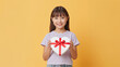 © bephoto - 黄色い背景でハート形の贈り物を持つ日本人の女の子の笑顔