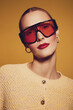 © SHOTPRIME STUDIO - Stylish woman with slicked back hair wears oversized red sunglasses and gold hoop earrings. She poses confidently in a textured yellow sweater against a warm orange background.