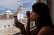 © Julien McRoberts - Young Latina woman applying makeup by window with church view in Mexico