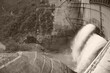 © BRUCE - 富山県の黒部ダムから力強く放水される観光放水の様子をセピア調で捉えた迫力ある風景。巨大なコンクリート構造物と周囲の険しい山々の対比が印象的な夏の日の眺めです。