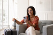 © fizkes - Smiling Brazilian woman using smartphone seated in armchair with suitcase while waits for taxi or airport transfer, prepare for flight, messaging through mobile app, make check-in, check boarding pass