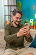 © Andrii Iemelianenko - Middle-aged man at home reads smartphone message smiles nodding with approval and joyful success. Young adult guy on sofa holds mobile phone enjoys positive notification showing cheery big confidence